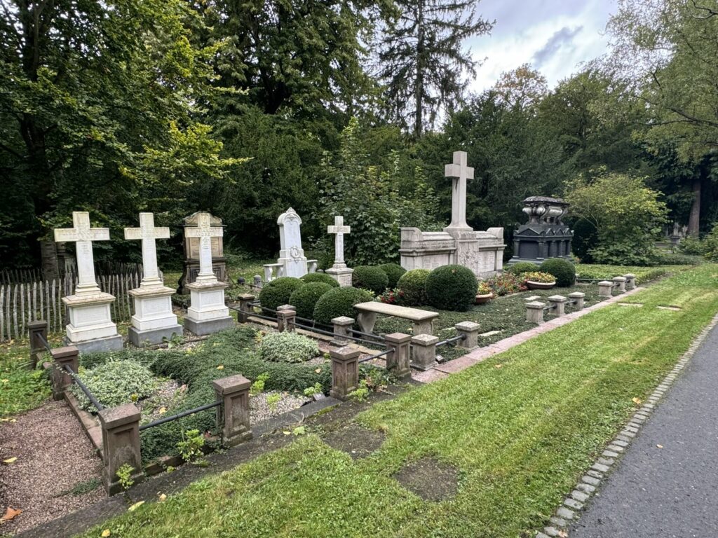 Ein gepflegter Friedhof mit mehreren Grabsteinen und Kreuzen, umgeben von grünen Büschen, Bäumen und einem grasbewachsenen Weg auf der rechten Seite unter einem bedeckten Himmel - typisch für Grabarten am Hauptfriedhof in Frankfurt am Main.