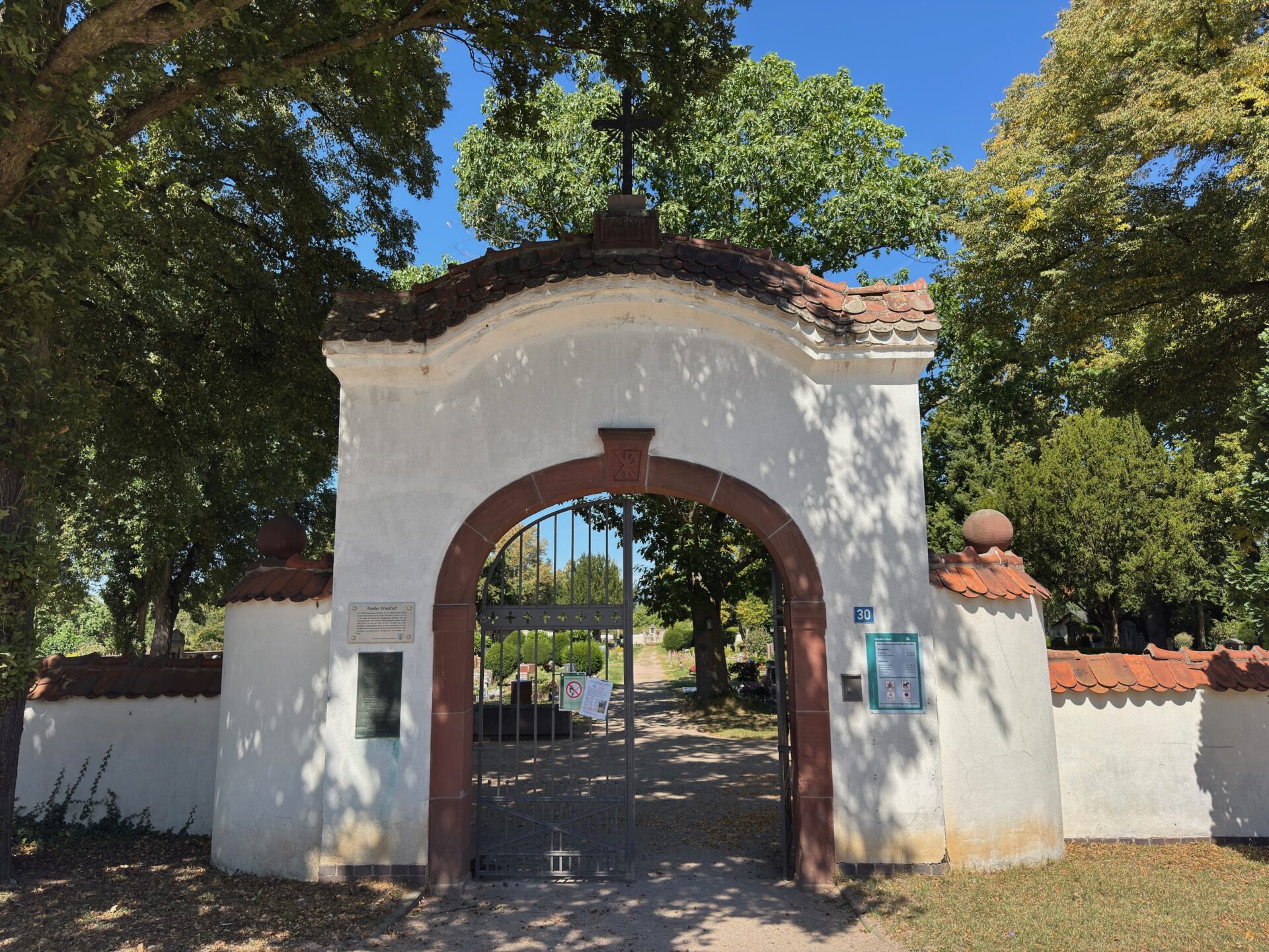 Der Friedhof Nied Frankfurt Eingang verfügt über ein weißes Bogentor mit Ziegeldach und Eisentor, das zu einem Friedhof führt, der von hohen grünen Bäumen unter einem klaren blauen Himmel eingerahmt wird. Zwei Hinweistafeln und eine Gedenktafel zieren die Wände des Tors.