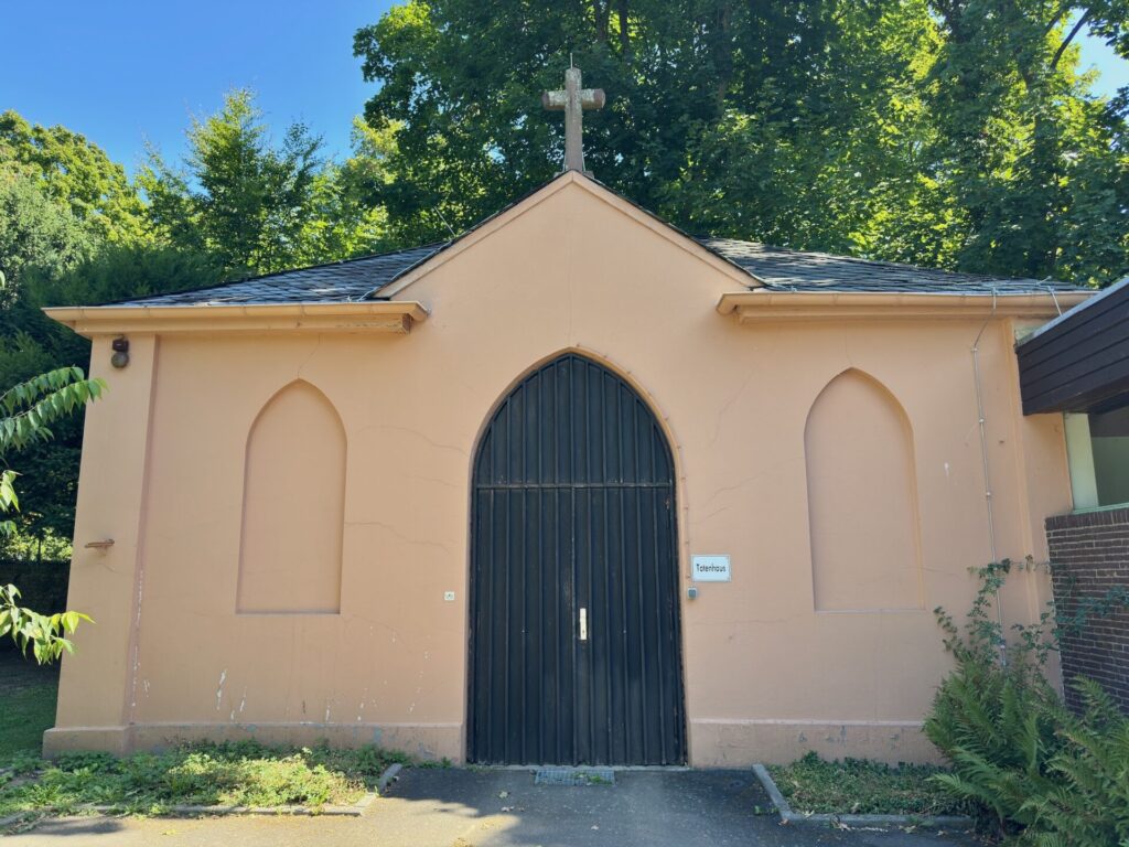 Auf dem Friedhof Sossenheim in Frankfurt am Main steht ein kleines, pfirsichfarbenes Gebäude, das sogenannte Totenhaus. Mit einem Kreuz auf dem Dach, bogenförmigen Fassadendetails und einer hohen schwarzen Bogentür ist es von grünen Bäumen und üppigen Pflanzen umgeben.