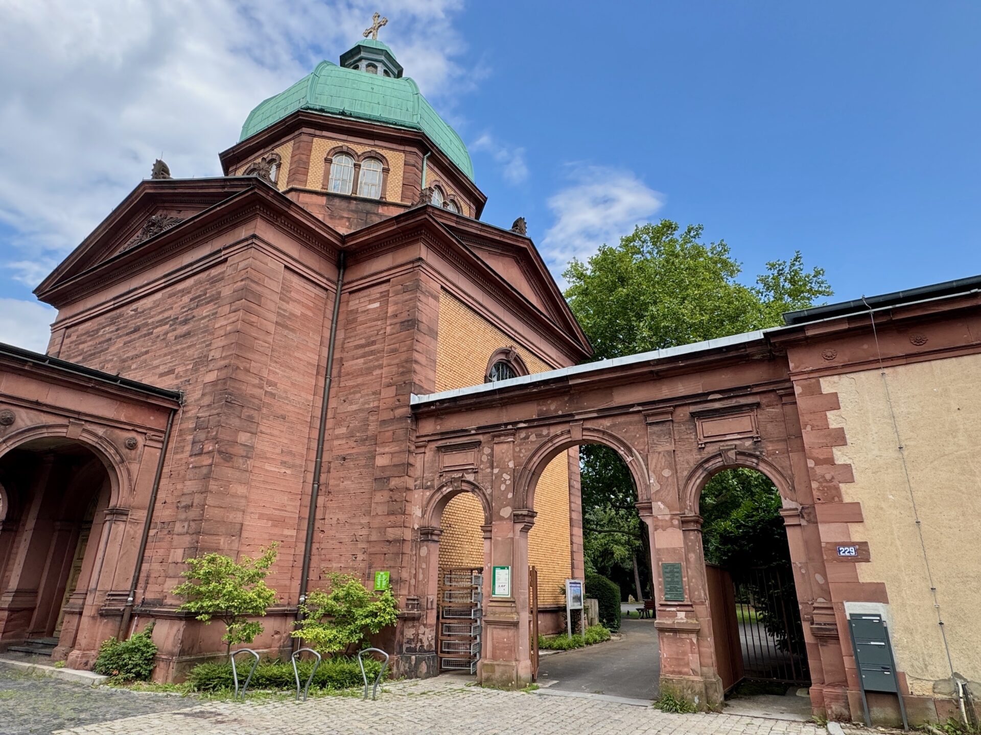 Die historische Trauerhalle, ein rotes Backsteingebäude mit grünem Kuppeldach und bogenförmigem Eingang aus Stein steht auf dem Gelände des Südfriedhofs Sachsenhausen in Frankfurt am Main, vor einem teilweise bewölkten Himmel, umgeben von Grünflächen und einem kopfsteingepflasterten Weg.